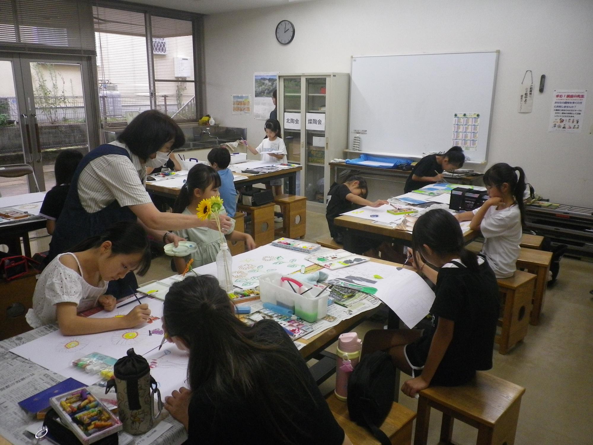 新聞紙を広げた机の上に水彩絵の具や筆が置かれ、花や自然を題材にした絵を描いており、教師が一人の子どもに花を見せながら指導している様子の写真