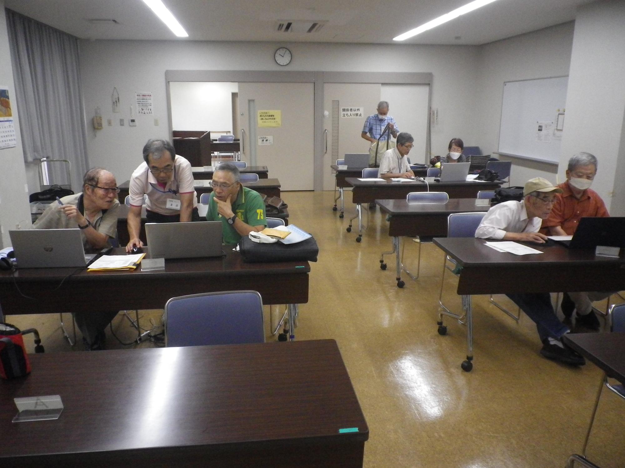 複数の参加者がパソコンの前に座り、前田昭枝さんが画面をのぞきこんで指導している、パソコン教室の写真