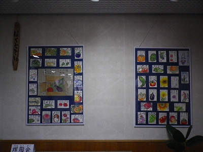 白い壁に、野菜や果物の絵が描かれたポストカードなどが多数配置されている青色の額が2つ掛けられている展示の様子の写真