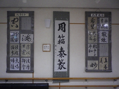 白い壁に、書道作品が貼られた薄いグレーの台紙が3つ掛けられており、中央には縦長の大きな作品が一枚、両側には様々な文字の書道作品が複数掲示されている展示の写真