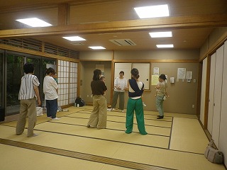 和室の畳の上で、数人の女性たちが子供を抱っこして立ち、講師から手遊びや子育てに関する指導を受けている様子の写真