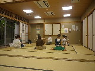 和室の畳の上で正面に講師が手遊びの見本を見せ、複数の親子が子供を膝に座らせたりしながら手遊びをしている様子の写真