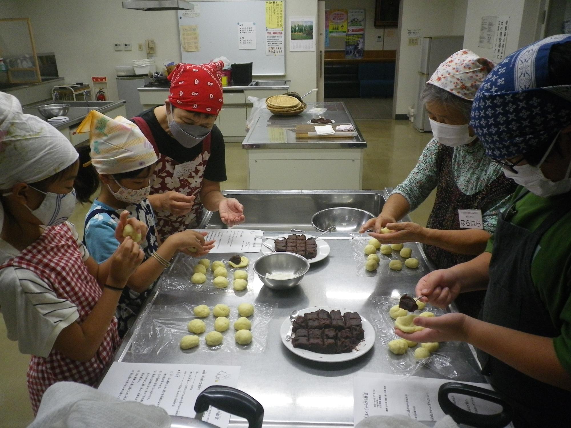 調理室のステンレス製テーブルを囲み、エプロンとバンダナ、マスクを着用した大人と子供が、あんこを黄色い生地で包んで丸める作業に熱中している、お月見まんじゅう作り教室の写真