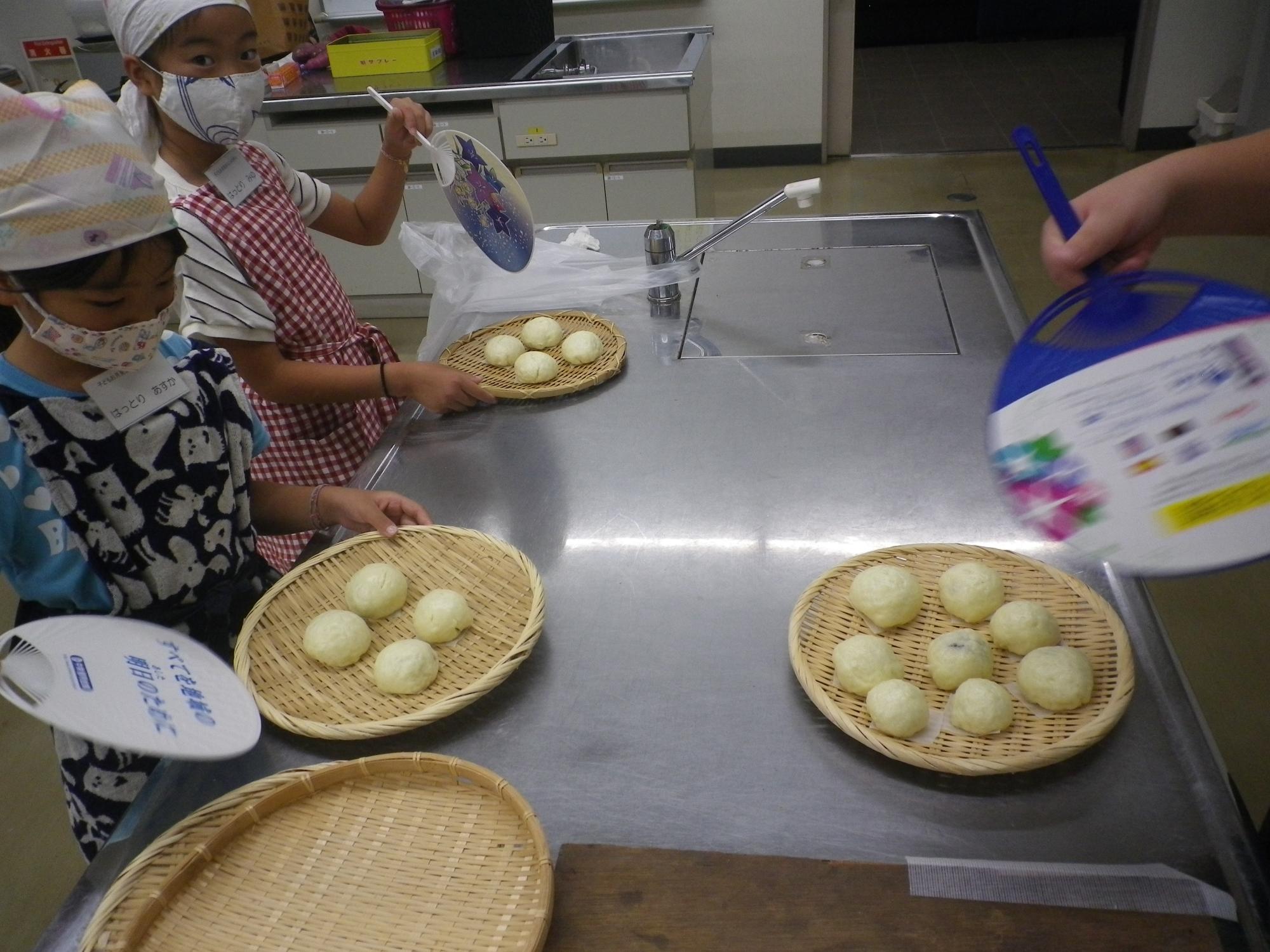 調理室のステンレス製調理台の上で、頭巾とマスクを着用した二人の子供が、丸く成形されたお月見まんじゅうを竹ざるに乗せて、うちわで扇いでいる様子の写真