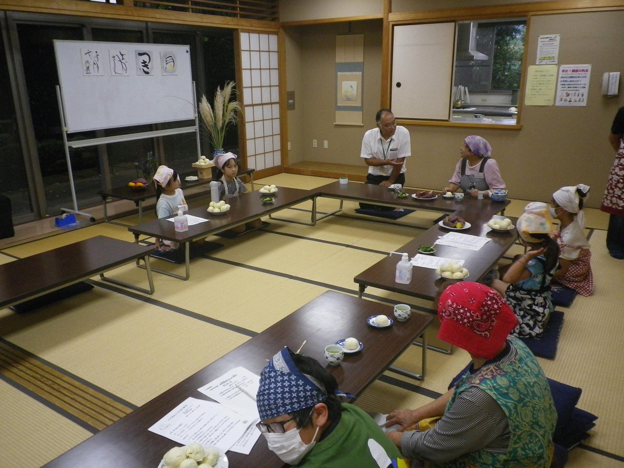 和室の広間で、数名の子供と大人が座卓を囲んで座り、それぞれ頭巾やバンダナを着用して、丸いお月見まんじゅうが乗った皿を見ながら、座って話しをしている様子の写真