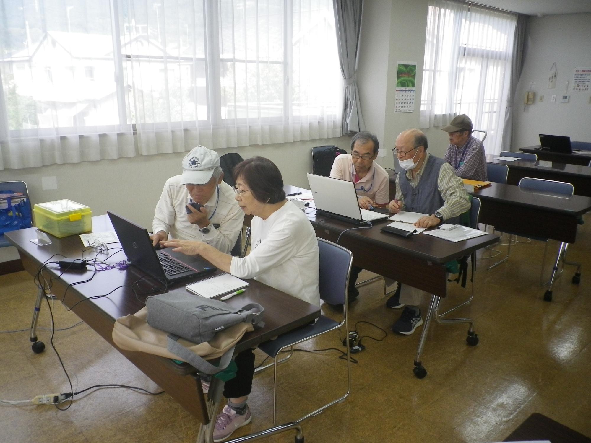 大きな窓から明るい光が差し込む室内の長机で、複数の参加者が各自のノートパソコンを操作し、教えてもらっているパソコン教室の写真