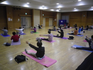 色とりどりのヨガマットの上に座り、両手、両足を伸ばしヨガをしている様子の写真