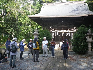 緑豊かな森の中にある社殿の前に、リュックや帽子を身に着けた参加者が、境内で説明を聞いている様子の写真