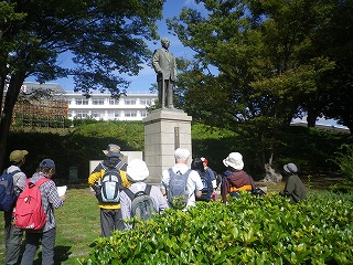 青空の下、緑に囲まれた大学で、リュックサックを背負った数名のグループが、台座に乗った偉人の銅像を取り囲み、何かの説明を聞いている様子の写真