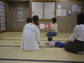 和室の畳の上で、女性が子どもたちに向けてウサギの絵が描かれた絵本を読み聞かせており、2組の親子が熱心に聞き入っている様子の写真