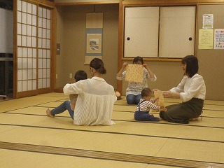 和室の畳の上で、2組の親子が乳幼児を抱いたり前に座らせたりしながら、柄物の布を使ってハンカチ遊びをしている様子の写真