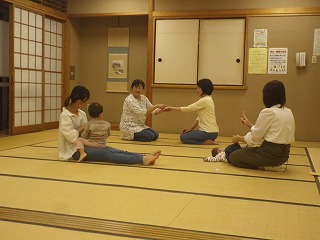 和室の畳の上で正面に講師が手遊びの見本を見せ、2組の親子が子供を膝に向かい合うように座らせたり、寝かせたりしながら手遊びをしている様子の写真