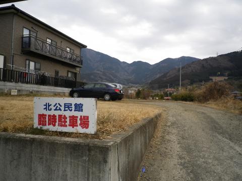 北公民館臨時駐車場の看板が立ち、奥に2階建ての住宅と山並みが見える第2駐車場の写真