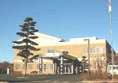 青空の下、ベージュ色の煉瓦のような外壁を持つ近代的な建物の正面玄関を捉えており、手前には立派な松の木と国旗がはためく高いポールが立っている北公民館外観写真