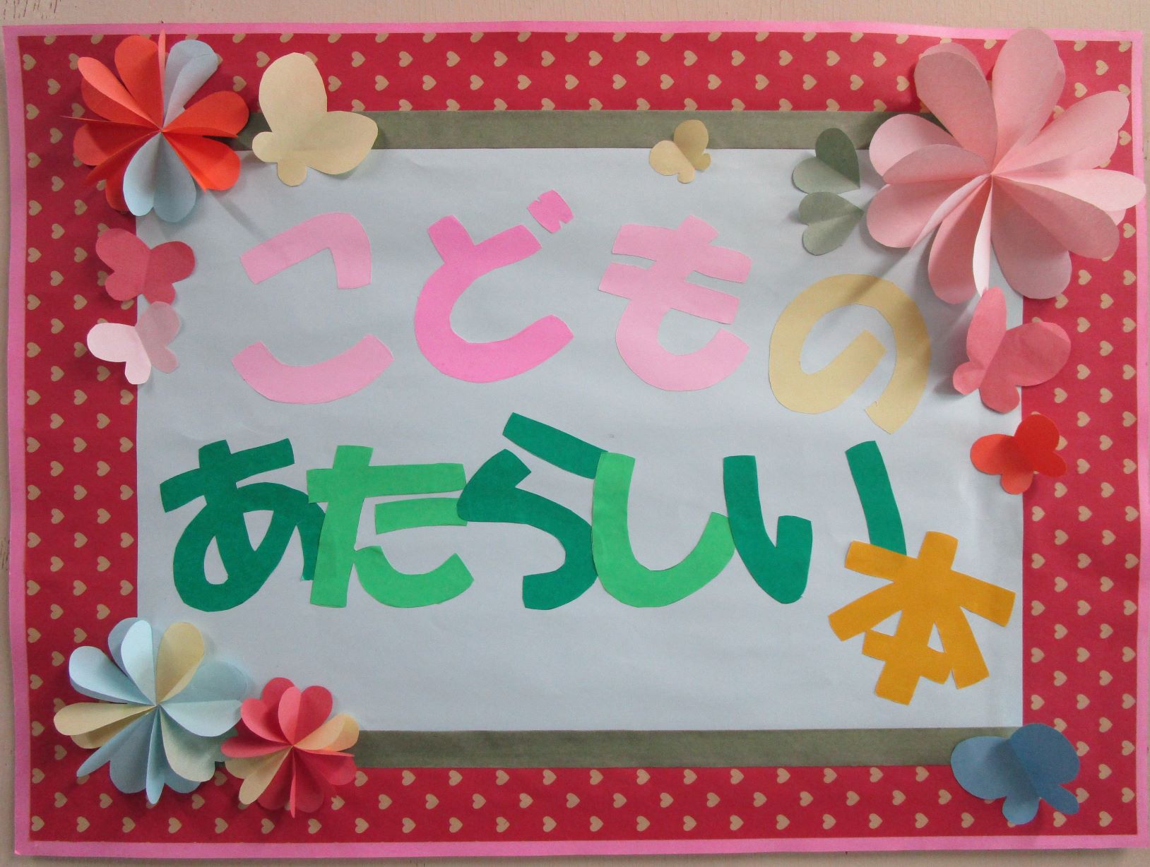 赤色のハート柄の枠で縁取られた掲示板に、色画用紙で切り抜かれた「こどものあたらしい本」の文字と、立体的な花や蝶の飾りが賑やかに配置された掲示物の写真