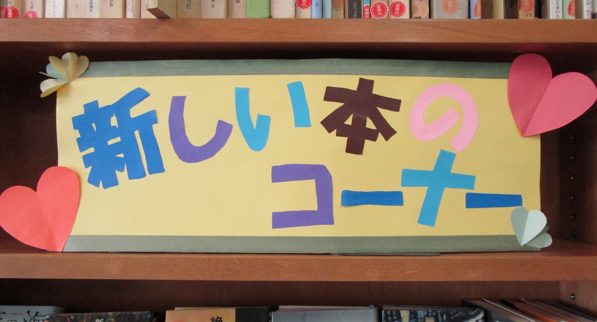 木製の本棚に、黄色の台紙に様々な色の切り文字で「新しい本のコーナー」と書かれ、折り紙でできたハートや葉が装飾されたポップが置かれている写真