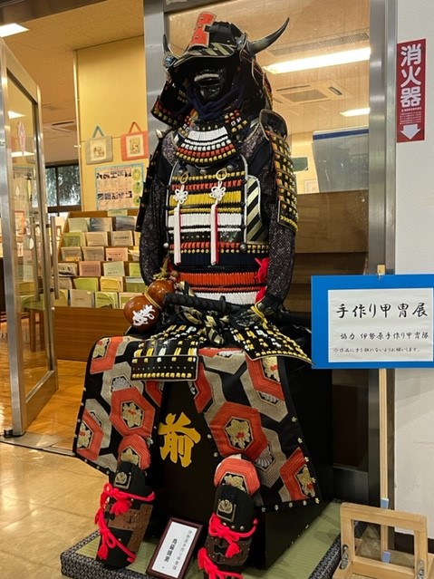 縁は朱色、胴体は黒や黄色、赤色などの様々な色と文様で飾られた、兜と陣羽織を身につけた華やかな甲冑が、台座に座った状態で展示されている写真