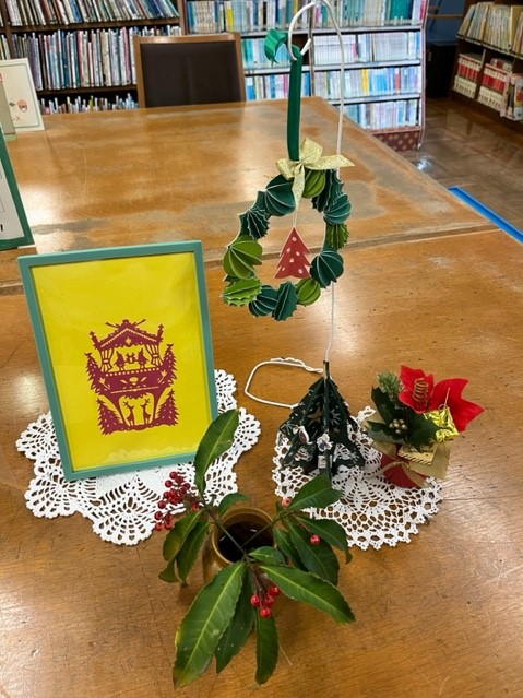 クリスマスリース、額に入った切り絵、ポインセチアの造花、赤い実のついた植物が白いレースの敷物の上に並べられた写真