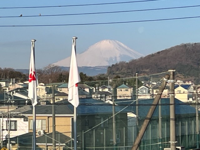 青空の下、手前に住宅地と校庭があり、2本の国旗掲揚ポールが立っている中、遠景に雪をかぶった富士山がそびえ立つ様子を捉えた写真