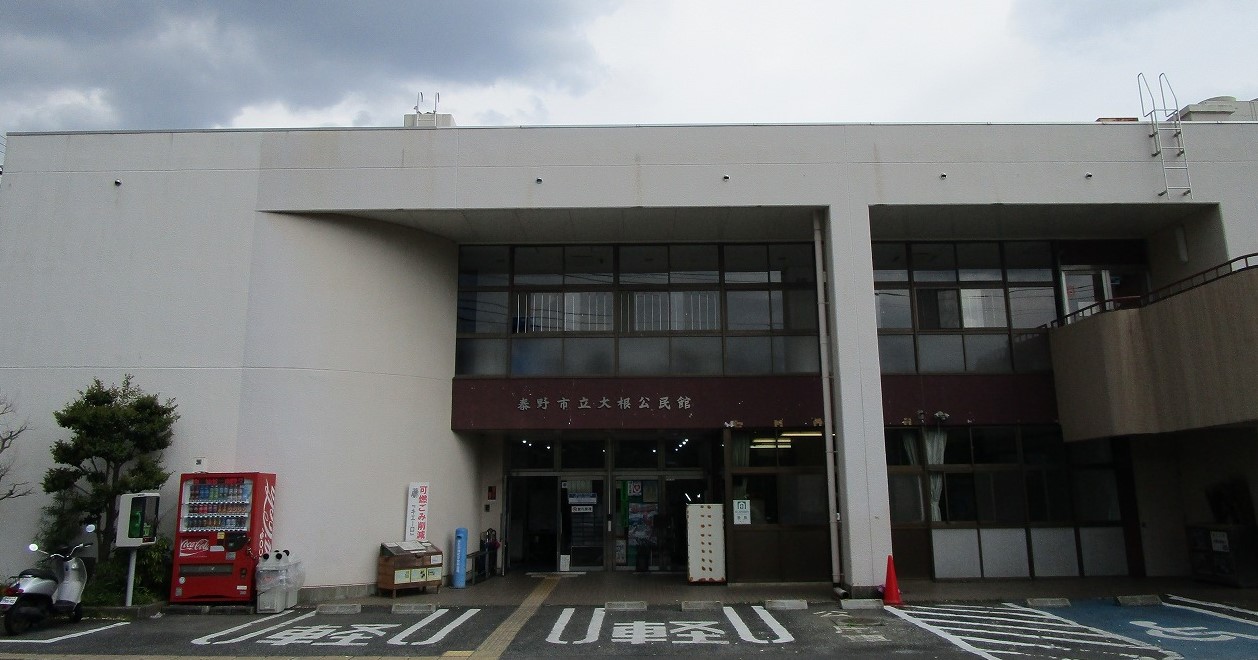 白い外壁の建物の正面を捉えており、中央の入り口の上には「秦野市立大根公民館」と書かれた看板があり、赤色の自動販売機などが脇に置かれている大根公民館の写真