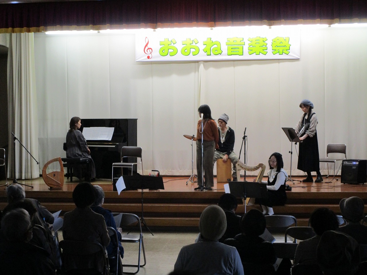 ピアノやハープ、打楽器などが置かれたステージ上で、複数の演奏者と歌い手が楽器の練習や打ち合わせをしている様子を観客席から撮影した写真