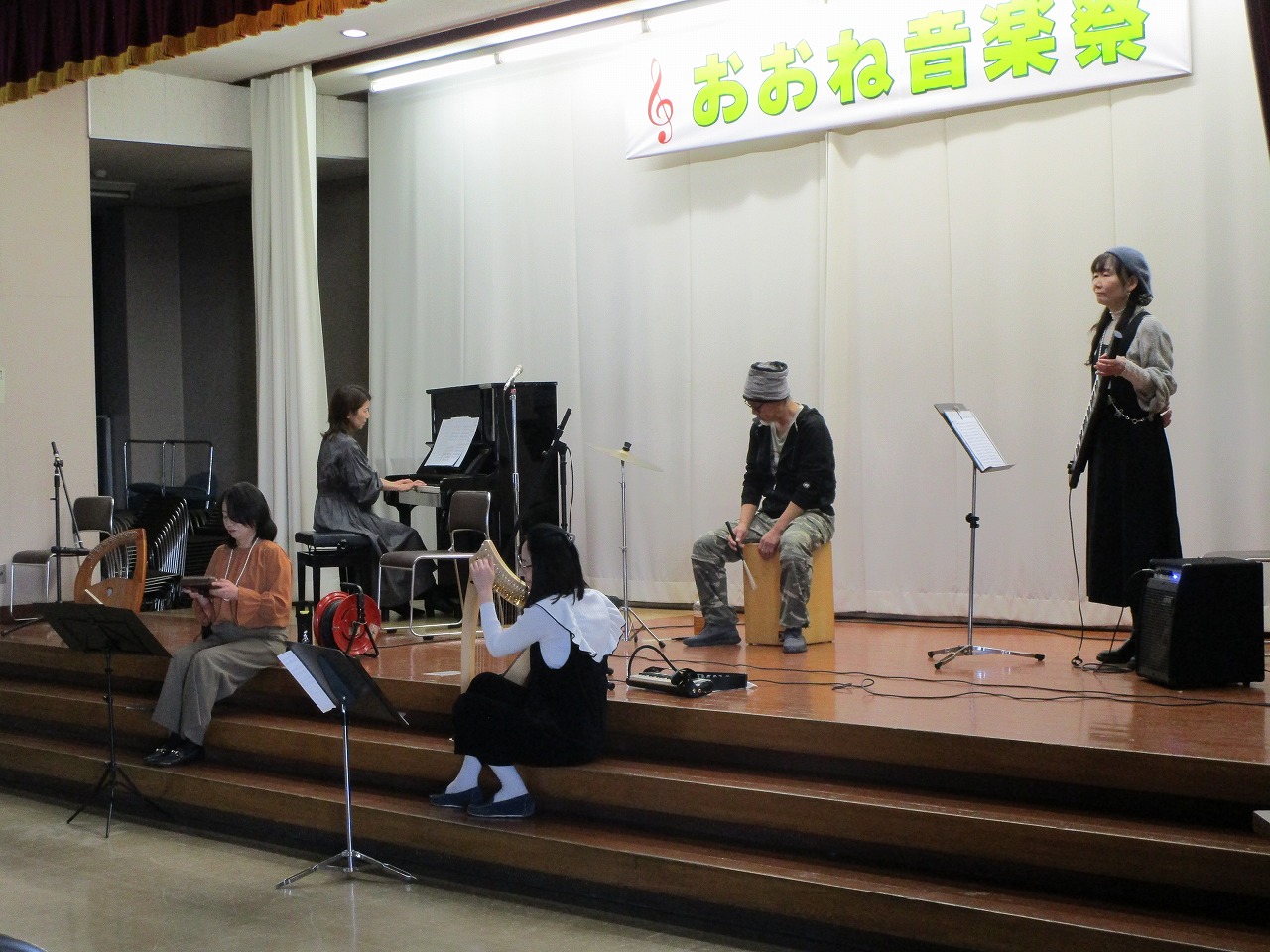 音楽祭のリハーサルで、舞台前の階段に座る人や舞台上でハープやピアノなどの楽器を演奏する人々を写した写真