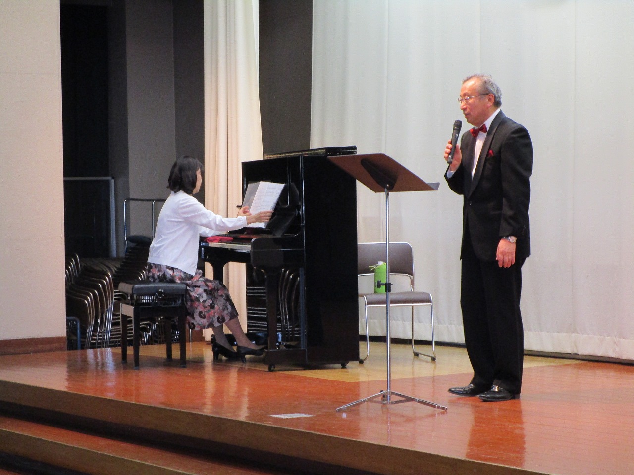 赤い蝶ネクタイとタキシードを着用した男性がマイクで歌を披露しており、その横で女性がピアノを弾いている、ステージでの演奏風景を写した写真