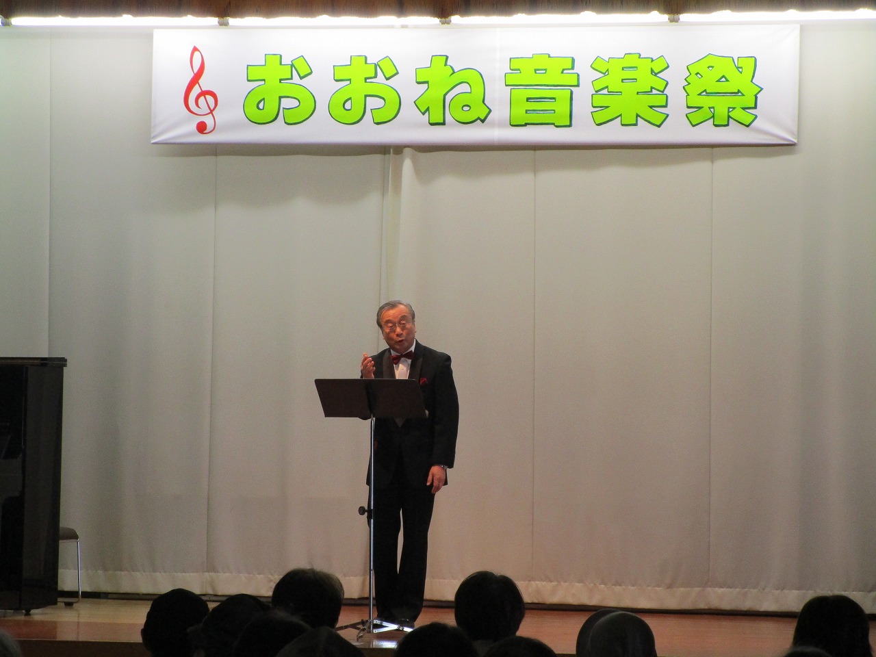 「おおね音楽祭」の横断幕が掲げられたステージ上で、タキシード姿の男性がマイクに向かって歌を披露している写真