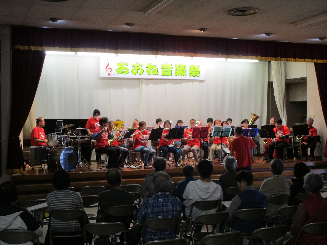 「おおね音楽祭」の横断幕が掲げられたステージ上で、赤いTシャツを着用した複数の演奏者たちが、ドラムや管楽器などを演奏している写真