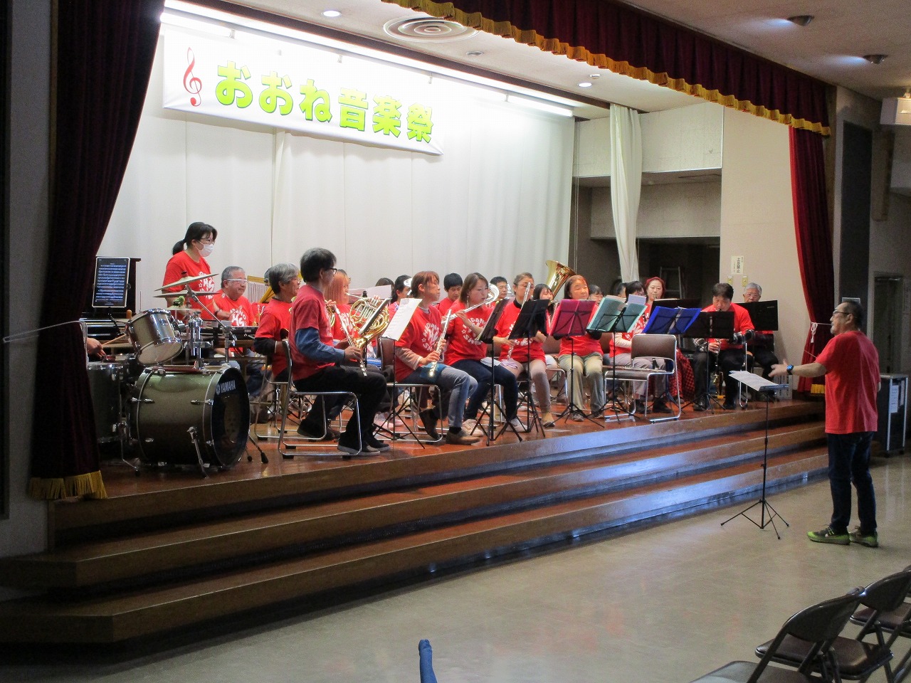 「おおね音楽祭」の横断幕が掲げられたステージ上で、赤いTシャツを着用した複数の演奏者が、指揮者のもと、ドラムや管楽器を演奏している写真