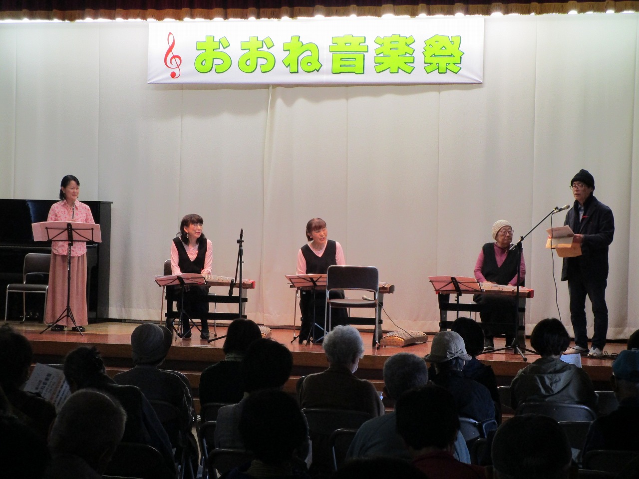 「おおね音楽祭」の横断幕が掲げられたステージ上で、5人の男女おり、男性がマイクスタンドで観客に向けて話をしている写真