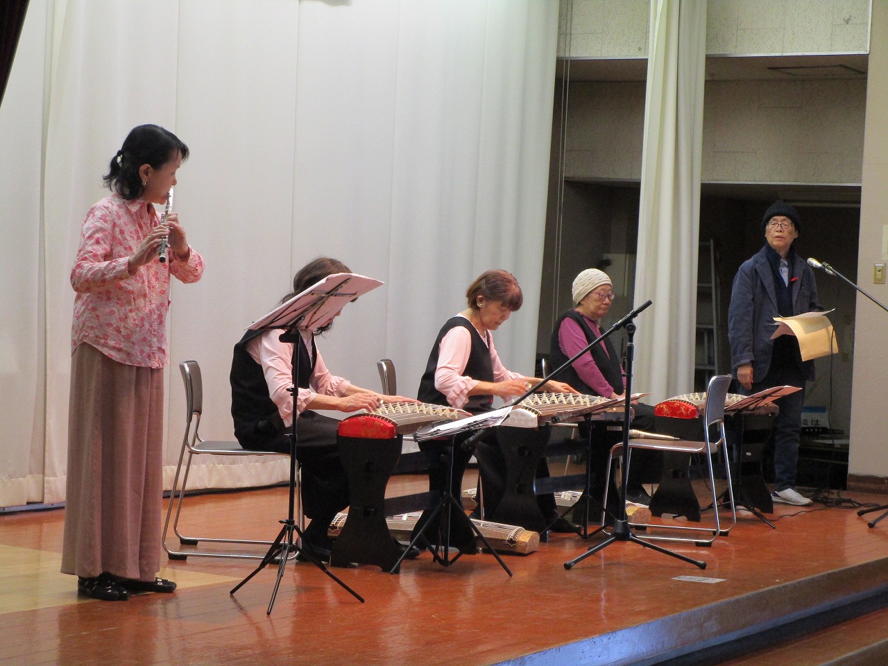 マイクや譜面台が設置されているステージ上で、数人の男女がフルートや大正琴などを演奏している写真