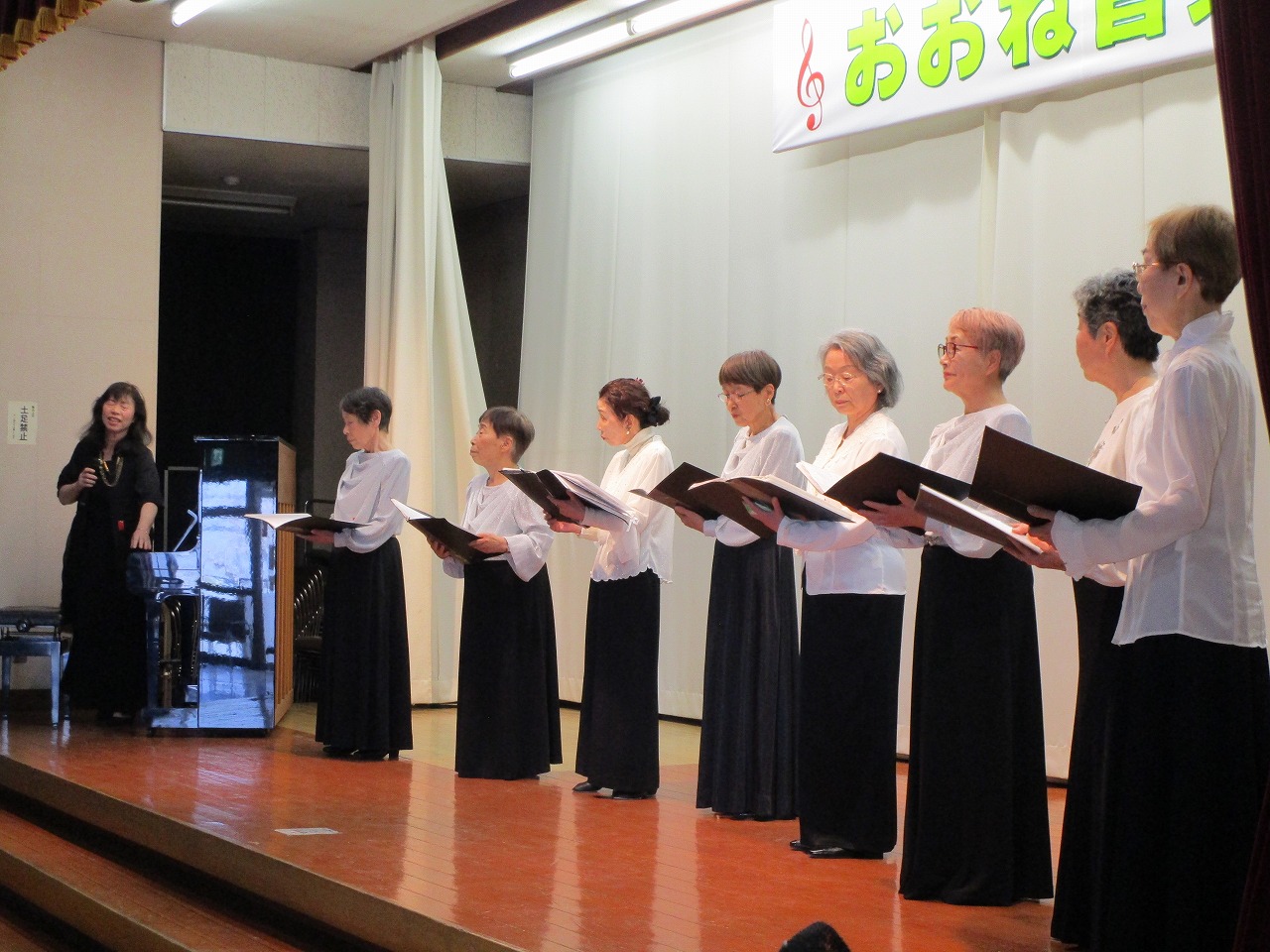 「おおね音楽祭」の横断幕が掲げられたステージ上で、楽譜を手に合唱している複数の女性たちの写真