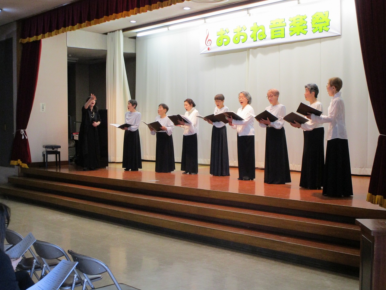 「おおね音楽祭」の横断幕が掲げられたステージ上で、黒いスカートと白いブラウスを着た複数の女性たちが、観客に向かって歌を披露している写真