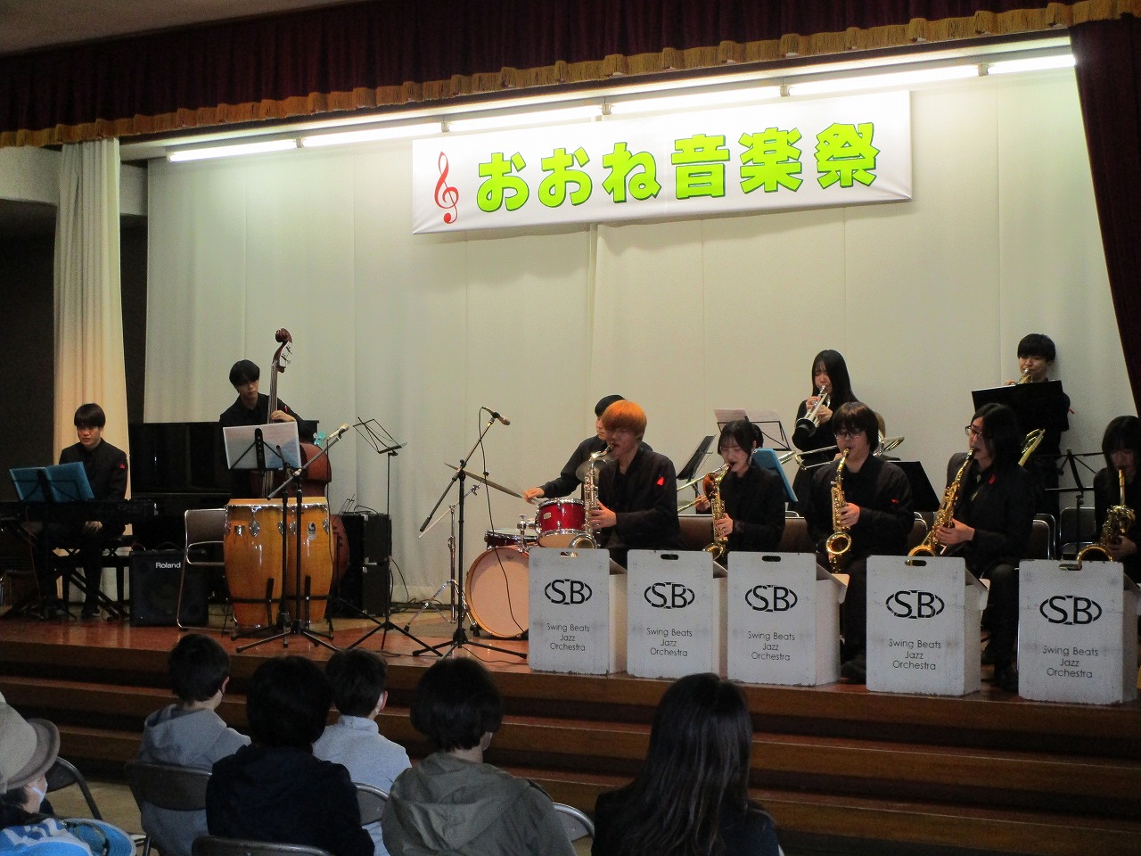 「おおね音楽祭」の横断幕が掲げられたステージ上で、複数の演奏者がドラムやベース、サックスなどの楽器を演奏している、ジャズバンドの写真