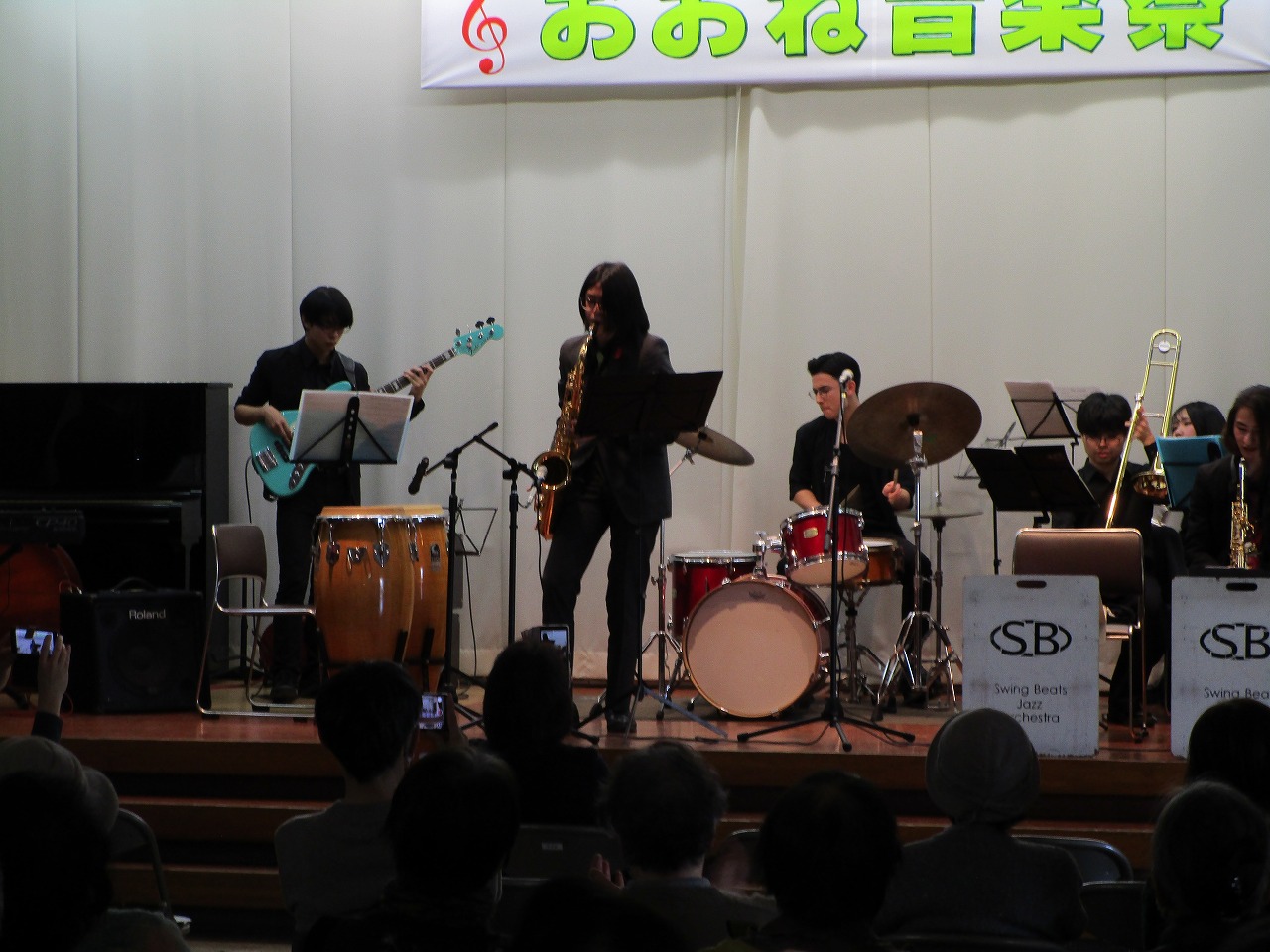 「おおね音楽祭」の横断幕が掲げられたステージ上で、複数の演奏者が楽器を演奏しているジャズバンドの中で、サックス演奏者が中央で立って演奏している写真