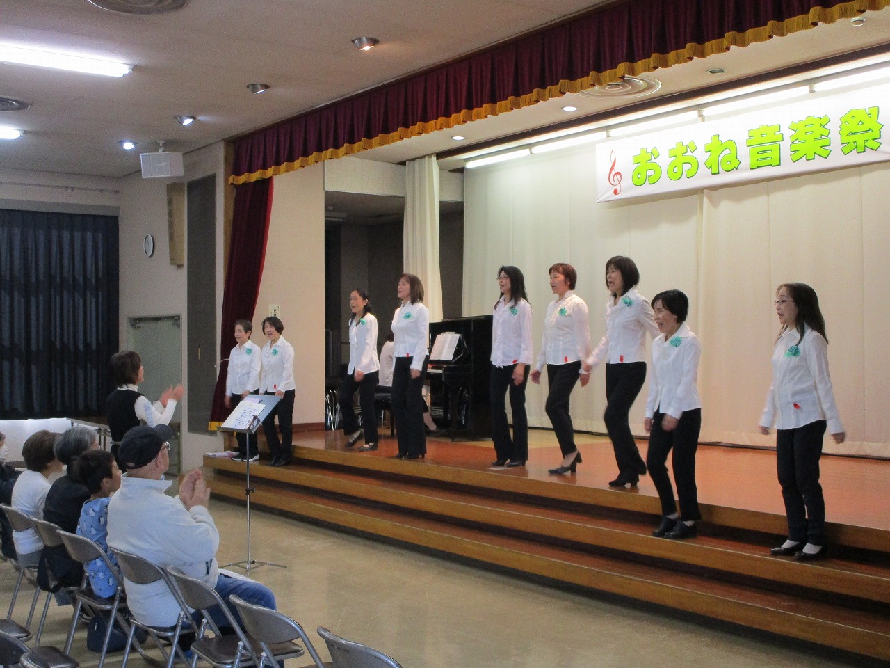 「おおね音楽祭」と書かれた横断幕が掲げられたステージ上で、白いシャツと黒いパンツを着用した複数の女性たちが、観客に向かって歌を披露しているところをステージ端から撮影した写真