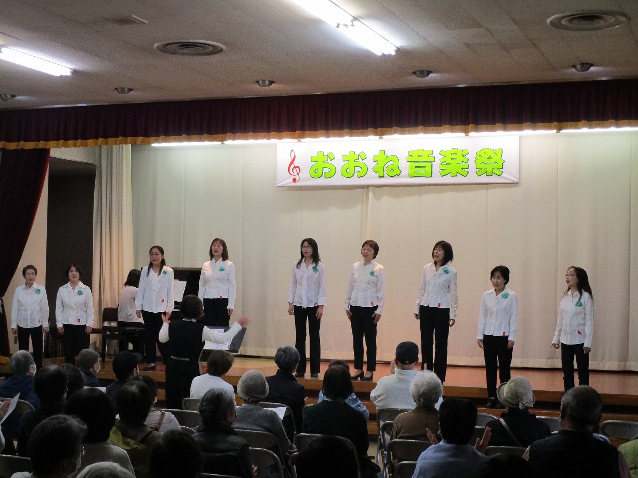 「おおね音楽祭」の横断幕が掲げられたステージ上で、白いシャツと黒いパンツを着用した複数の女性たちが、観客に向かって歌を披露しているところを客席後方から撮影した写真