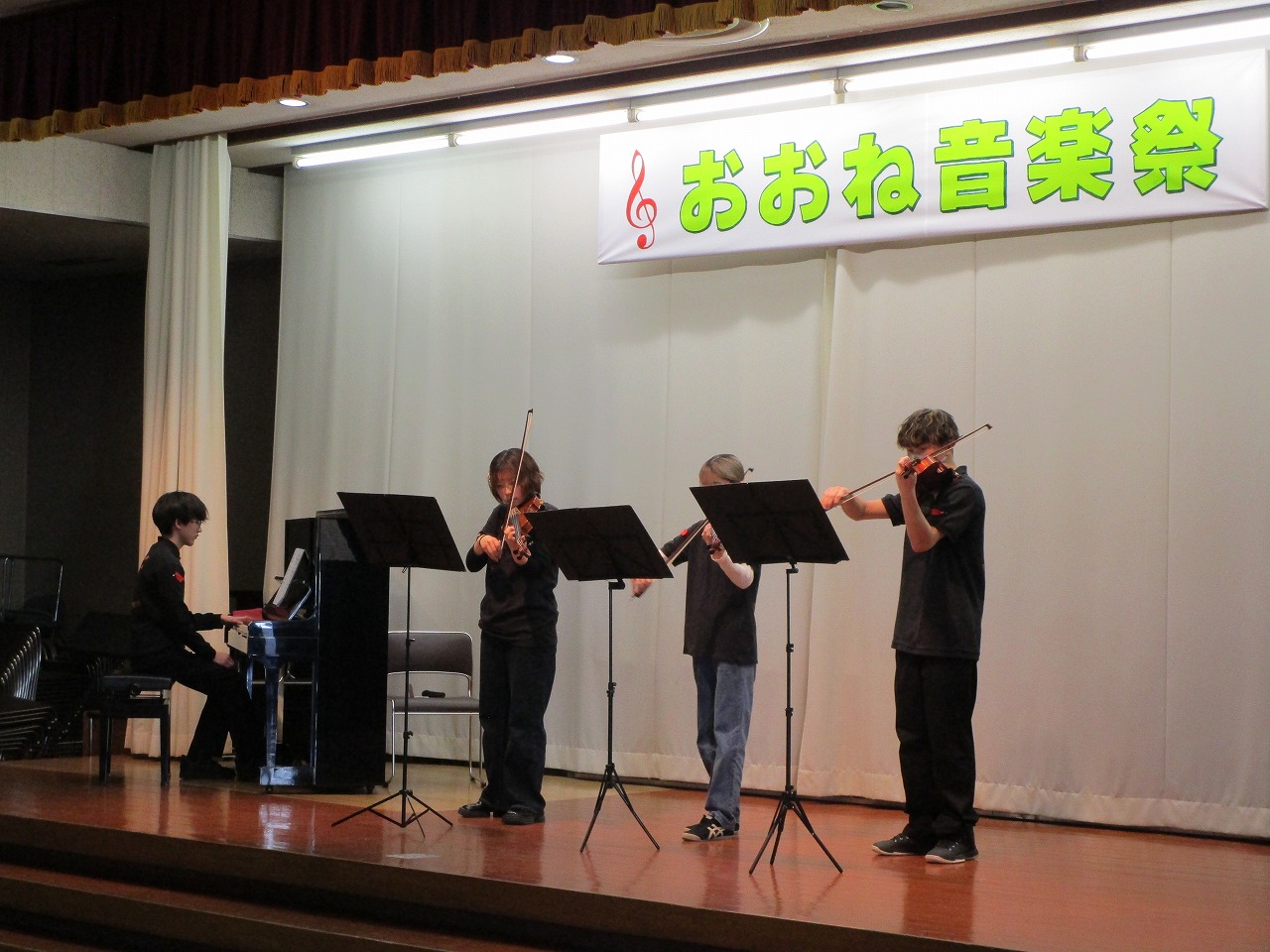 黒いシャツとズボンを着用した4人の男女が、ステージ上でピアノとヴァイオリンを演奏している写真