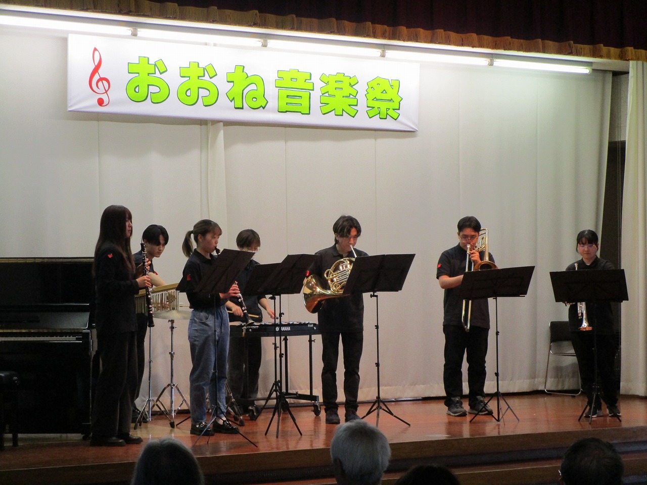 「おおね音楽祭」の横断幕が掲げられたステージ上で、複数の演奏者がトランペットやトロンボーンなどの楽器を演奏している写真