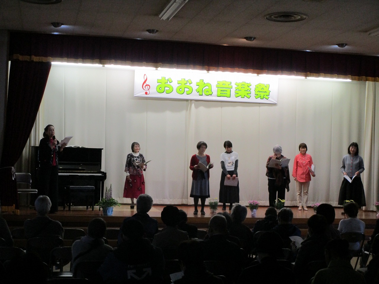 「おおね音楽祭」の横断幕が掲げられ、指揮者やピアノ奏者を含む複数の女性たちが観客の前で演奏をしている写真