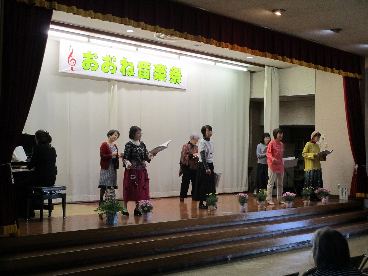 「おおね音楽祭」と書かれた横断幕が掲げられたステージ上で、複数の大人がステージ上で合唱をしている写真