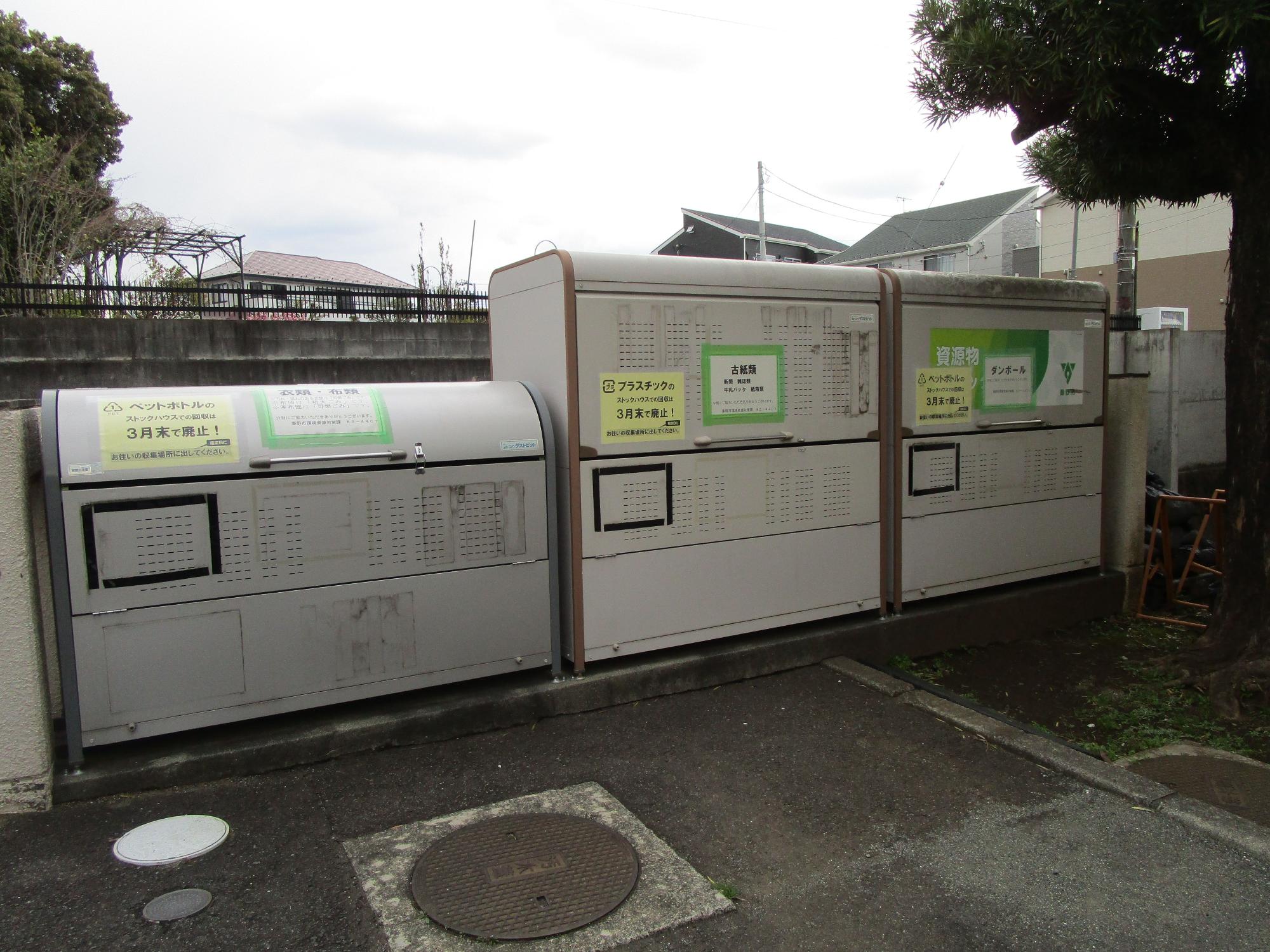 住宅街の空き地または敷地の一角に、ペットボトル、プラスチック、古紙と空き缶など、資源ごみの種類ごとに投入口と説明ラベルが設けられた大型の資源回収ボックスが三台並んでいる屋外の写真