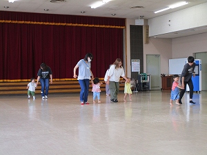 ホールの舞台前にある広くて明るい床の上で、数人の母親らしき大人が幼児の手を引いて歩く練習や遊びを行っている写真