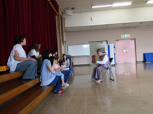 赤い緞帳が引かれた舞台下の階段に数人の母親と幼児が座り、向かい側の椅子に座った女性が絵本を読んで聞かせている写真