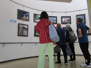 曲線を描く白い壁面のギャラリーで、参加者の来場者が、展示されている様々な風景を収めた額装写真を熱心に鑑賞している様子の写真