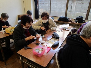 明るい窓際に面した室内で、マスクを着用した女性たちが机を囲み、裁縫道具を用いて和やかに手芸や手作業に取り組んでいる様子の写真