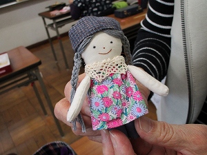 布製で、花柄の服を着て、グレーの三つ編みとチェックの帽子を被った、愛らしい表情の手作りの人形を大人が両手で持ち上げている様子の写真