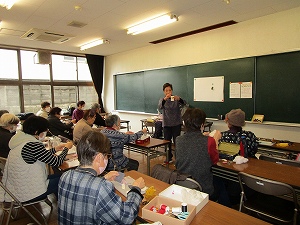 高齢者が集まった室内で、女性が黒板の前で何かを手に持ち説明しており、参加者は机に向かい手芸作業をしている様子の写真