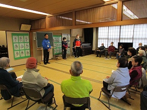 和室で高齢者たちが椅子に座って環状に集まり、正面に立つ三人の人物が参加者へ向けて何かを説明している様子の写真